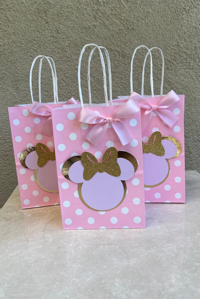 May include: Three pink polka dot gift bags with white handles and pink bows. Each bag features a Minnie Mouse design with a gold glitter bow and gold outline. The bags are for a party.