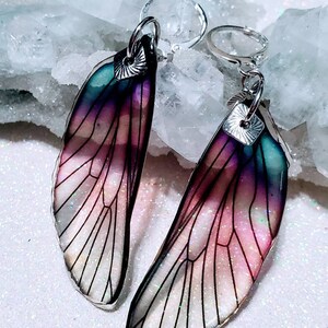 May include: A pair of iridescent fairy wing earrings. The wings feature a gradient of colors, including teal, purple, and pink, with black vein detailing. The earrings have silver-colored lever-back closures.