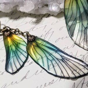 May include: A pair of handcrafted earrings featuring iridescent, translucent fairy wings. The wings transition from yellow to green and blue, with delicate black vein detailing. The earrings have antique bronze-colored metal findings.