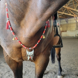 May include: A red beaded horse necklace with silver accents, tassels, and small bells. The necklace features a silver crescent moon pendant with feather and bead details. The horse is brown.