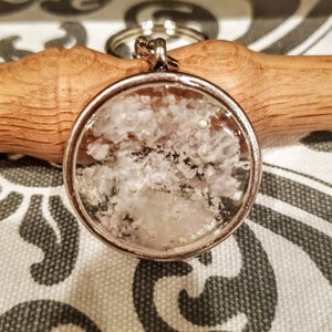 May include: A silver keychain with a round, clear resin charm containing white and silver glitter. The charm is attached to a silver key ring.