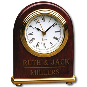 Peut inclure: Une horloge de bureau en bois d'acajou avec une bordure dorée et un cadran à chiffres romains. L'horloge porte une inscription personnalisée qui dit "Ruth & Jack Millers".