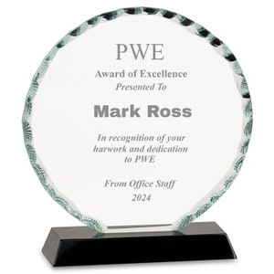 May include: A clear glass award with a black base. The award has a scalloped edge and features the text "PWE Award of Excellence Presented To Mark Ross In recognition of your harwork and dedication to PWE From Office Staff 2024".