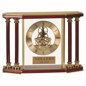 May include: A decorative clock with a gold-toned face and visible gears. The clock is encased in a rectangular frame with dark red wood and gold accents. The clock face features Roman numerals and the text "MILLERS EST 2017".