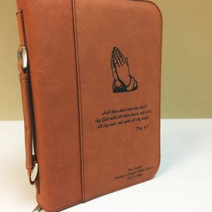 以下が含まれることがあります： ジッパー留めとシルバーのハンドルが付いたブラウンの革製の聖書カバー。カバーには、祈る手の彫刻と聖書の言葉「主なるあなたの神を、あなたの心とあなたの魂とあなたの力をもって愛しなさい。申命記 6:5」が刻まれています。カバーには、「Ron Smith Parkers Chapel FWB Church SD 01.1992」というテキストも含まれています。