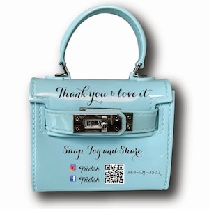May include: A light blue mini handbag with a silver lock and handle. The bag has the text "Thank you & love it" and "Snap. Tag and Share" printed on it. It also has a QR code and social media icons for Instagram and Facebook with the handle "Triedish".