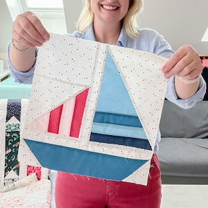 May include: A woman holds up a handmade quilt block featuring a sailboat design. The block is made of various shades of blue, pink, and white fabric. The background fabric is white with red polka dots.