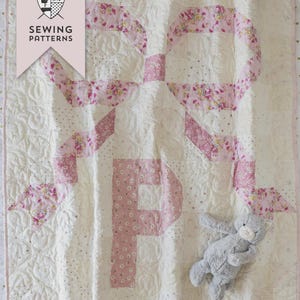 May include: A quilted baby blanket with a large pink letter "P" design. The quilt features a white background with pink floral accents and a gray stuffed animal. The text "BOWS & MONOGRAMS QUILT" is visible.