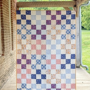 May include: A handmade quilt with a patchwork design. The quilt features a grid pattern of squares in various colors and patterns, including blue, pink, purple, and floral prints. The quilt is displayed outdoors, hanging from a wooden beam.