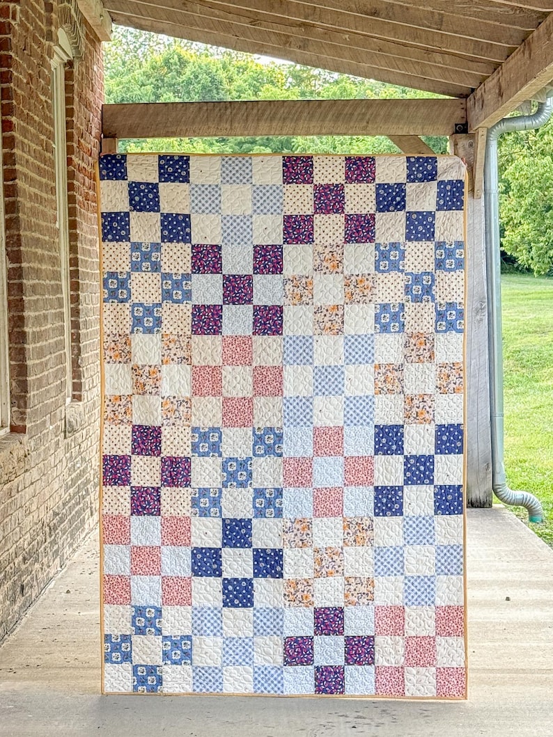 May include: A handmade quilt with a patchwork design. The quilt features a grid pattern of squares in various colors and patterns, including blue, pink, purple, and floral prints. The quilt is displayed outdoors, hanging from a wooden beam.