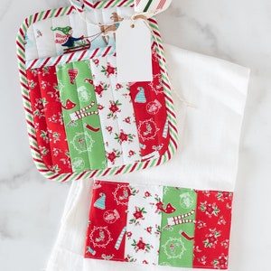 May include: A white kitchen towel with a red and green Christmas-themed fabric strip sewn on the bottom. A matching red and green pot holder with a Christmas-themed fabric design is on top of the towel. A white gift tag is tied to the pot holder with twine.