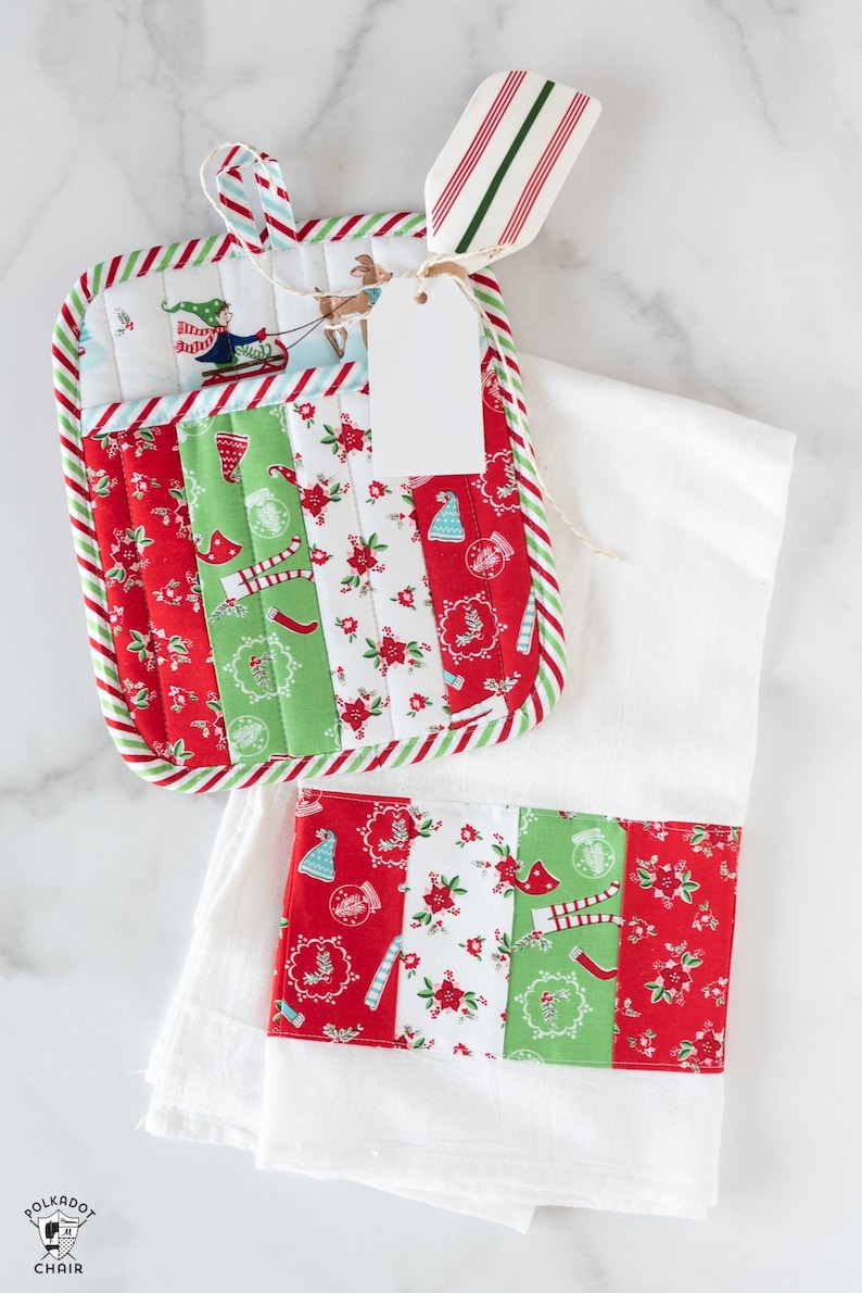May include: A white kitchen towel with a red and green Christmas-themed fabric strip sewn on the bottom. A matching red and green pot holder with a Christmas-themed fabric design is on top of the towel. A white gift tag is tied to the pot holder with twine.