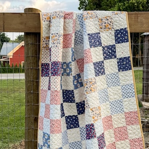 May include: A handmade patchwork quilt draped over a wooden fence. The quilt features a variety of patterned squares in shades of blue, red, and white, with a yellow border. The quilt is likely made of cotton and is a traditional style.