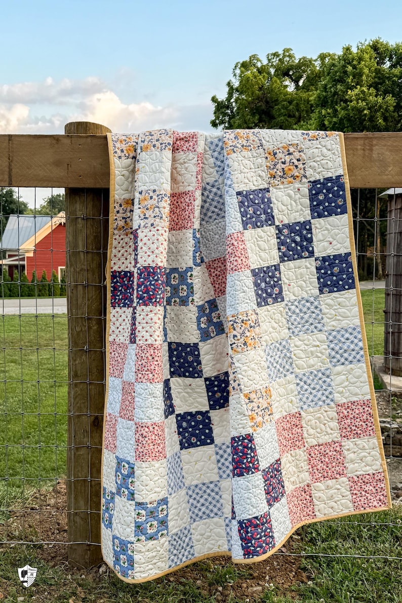 May include: A handmade patchwork quilt draped over a wooden fence. The quilt features a variety of patterned squares in shades of blue, red, and white, with a yellow border. The quilt is likely made of cotton and is a traditional style.