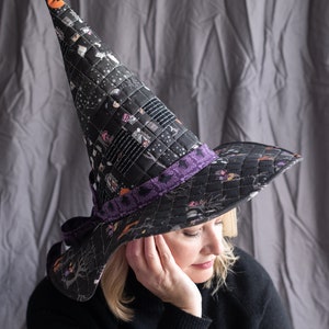 May include: A black witch hat with a purple ribbon and a print of houses and other Halloween imagery. The hat is worn by a person with blonde hair.