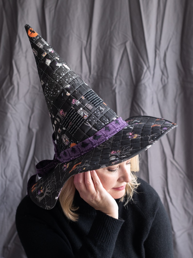 May include: A black witch hat with a purple ribbon and a print of houses and other Halloween imagery. The hat is worn by a person with blonde hair.