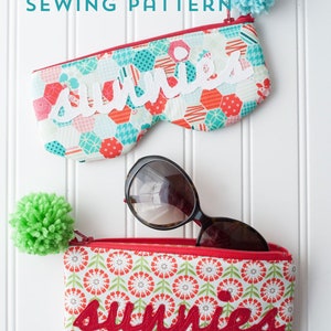 May include: Two colourful zippered pouches with the word "sunnies" embroidered on them. The pouches are made of fabric with different patterns. One pouch is turquoise and red with white embroidery, the other is red and green with red embroidery. Both pouches have pom-poms attached to the zipper pulls. Sunglasses are sitting on top of the red and green pouch.