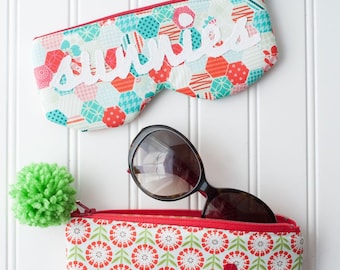 Puede incluir: Dos estuches con cremallera de colores con la palabra "sunnies" bordada. Los estuches están hechos de tela con diferentes patrones. Un estuche es turquesa y rojo con bordado blanco, el otro es rojo y verde con bordado rojo. Ambos estuches tienen pompones unidos a los tiradores de las cremalleras. Unas gafas de sol están encima del estuche rojo y verde.
