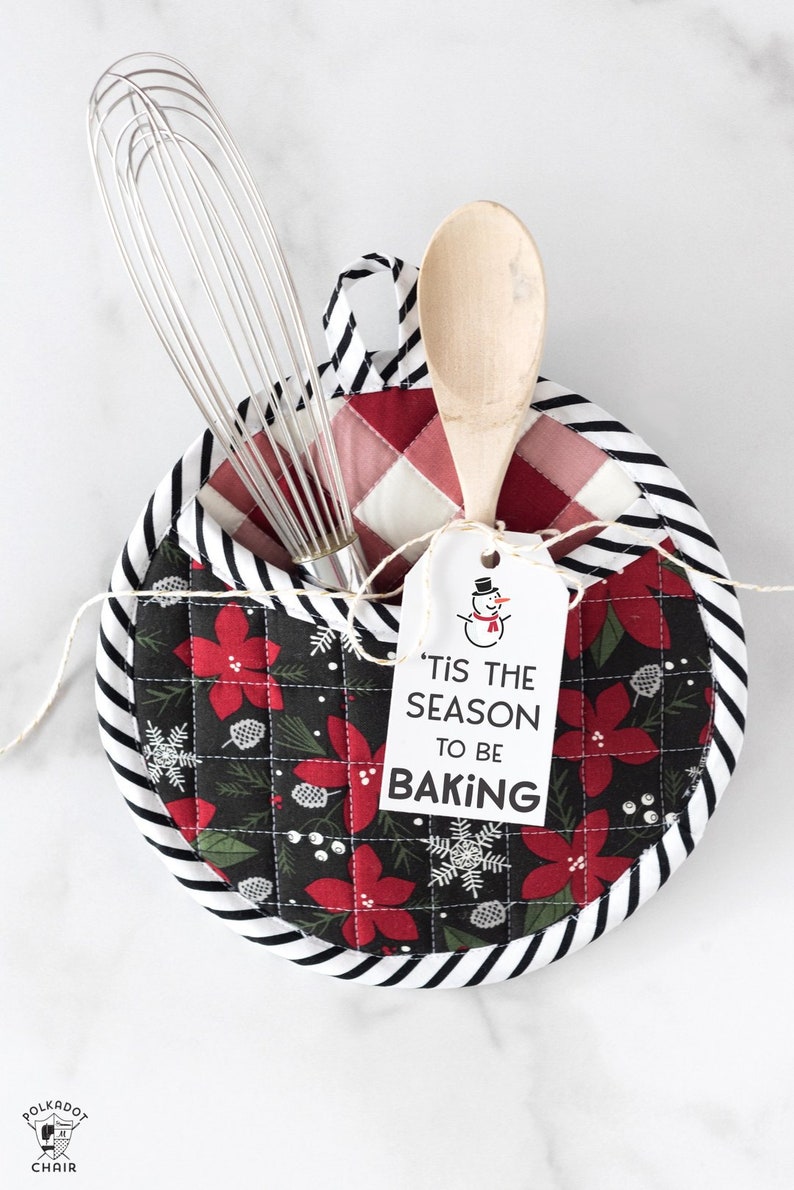 May include: A black and white striped pot holder with a red and green floral print. The pot holder has a pocket for utensils and a tag that says "'Tis the season to be baking" with a snowman illustration.