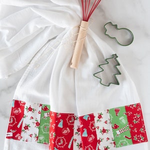 May include: A white kitchen towel with a red and green patchwork border. The towel is tied with twine and a red whisk. There are two green metal cookie cutters, one shaped like a snowman and the other like a Christmas tree.