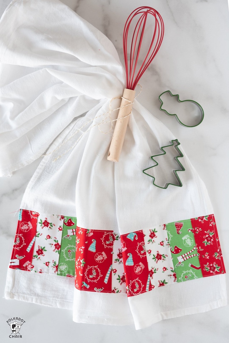 May include: A white kitchen towel with a red and green patchwork border. The towel is tied with twine and a red whisk. There are two green metal cookie cutters, one shaped like a snowman and the other like a Christmas tree.