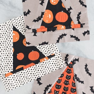 Puede incluir: Tres cuadrados de tela con diferentes patrones de temática de Halloween. El cuadrado superior tiene un fondo gris con murciélagos negros y un triángulo negro con calabazas naranjas. El cuadrado del medio tiene un fondo negro con calabazas naranjas y un fondo blanco con lunares negros. El cuadrado inferior tiene un fondo gris con murciélagos negros y un triángulo naranja con gatos negros.