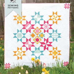 Op de afbeelding: Een quiltpatroon genaamd "Choose Happiness" met een witte achtergrond en een kleurrijk sterrendesign in tinten roze, geel, oranje en blauw. De quilt wordt getoond in een tuinsetting met groen gras en gele bloemen.