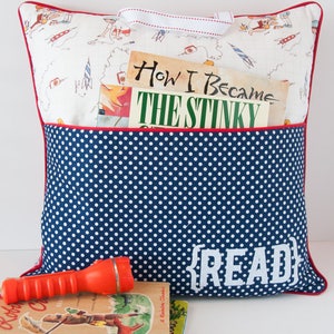May include: A blue and white polka dot pillow with a red trim and a pocket for books. The pillow has the word "READ" embroidered on the front. There are three books and a torch next to the pillow.