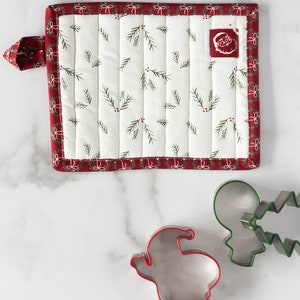 May include: A white quilted pot holder with a red border and a small Santa Claus applique. The pot holder has a loop for hanging. There are three cookie cutters on a white marble surface: a gingerbread man, a Christmas tree, and a Santa Claus.