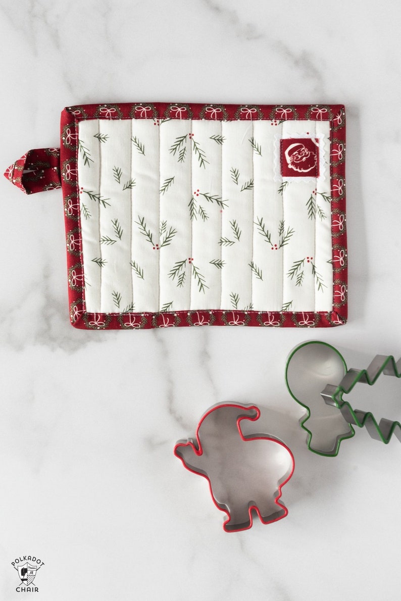 May include: A white quilted pot holder with a red border and a small Santa Claus applique. The pot holder has a loop for hanging. There are three cookie cutters on a white marble surface: a gingerbread man, a Christmas tree, and a Santa Claus.