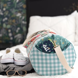 May include: A turquoise and white gingham patterned duffel bag with a patchwork design featuring a green and blue polka dot fabric, a floral print, and a geometric pattern. The bag has a cream-colored strap and a gold zipper pull.