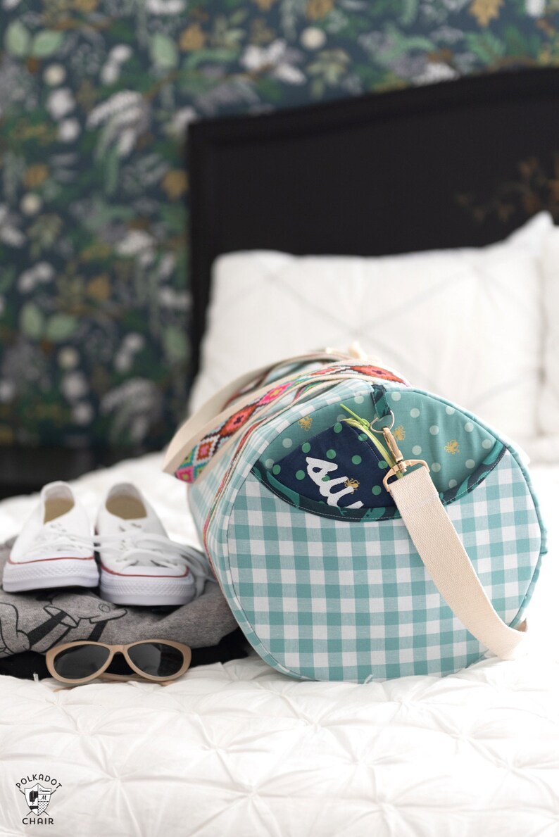 May include: A turquoise and white gingham patterned duffel bag with a patchwork design featuring a green and blue polka dot fabric, a floral print, and a geometric pattern. The bag has a cream-colored strap and a gold zipper pull.