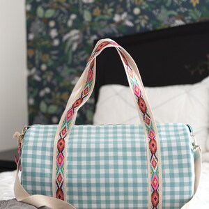 May include: A light blue and white gingham duffel bag with a white canvas strap and a colorful geometric patterned trim. The bag is sitting on a white bedspread.
