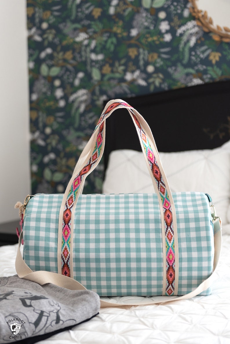 May include: A light blue and white gingham duffel bag with a white canvas strap and a colorful geometric patterned trim. The bag is sitting on a white bedspread.