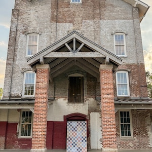 May include: Exterior view of a brick building with a gabled entrance and multiple windows. The building's facade is a mix of red and white bricks, with a quilt hanging in the doorway. The building has a chimney and a porch.