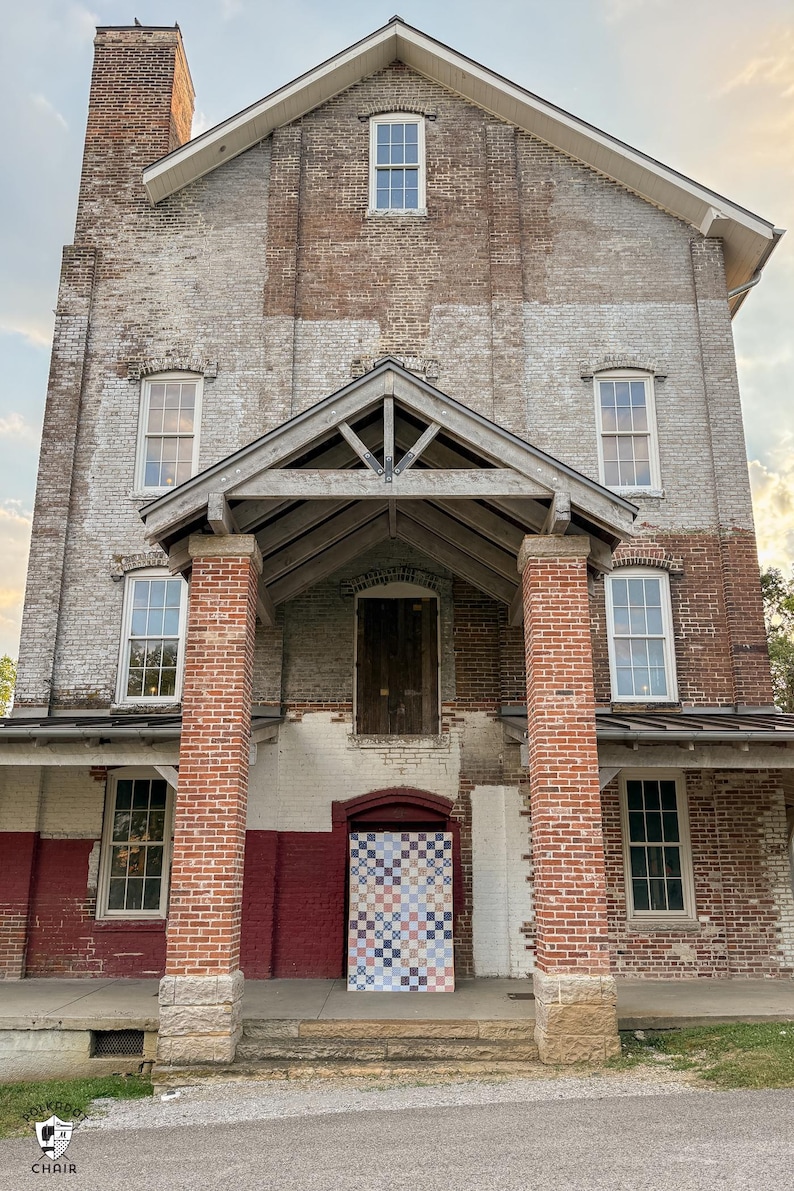 May include: Exterior view of a brick building with a gabled entrance and multiple windows. The building's facade is a mix of red and white bricks, with a quilt hanging in the doorway. The building has a chimney and a porch.