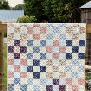 May include: A handmade quilt with a patchwork design. The quilt features a grid pattern of squares in various floral and solid prints, including pink, blue, and navy. The quilt is displayed outdoors, draped over a wooden fence, with a yellow border.
