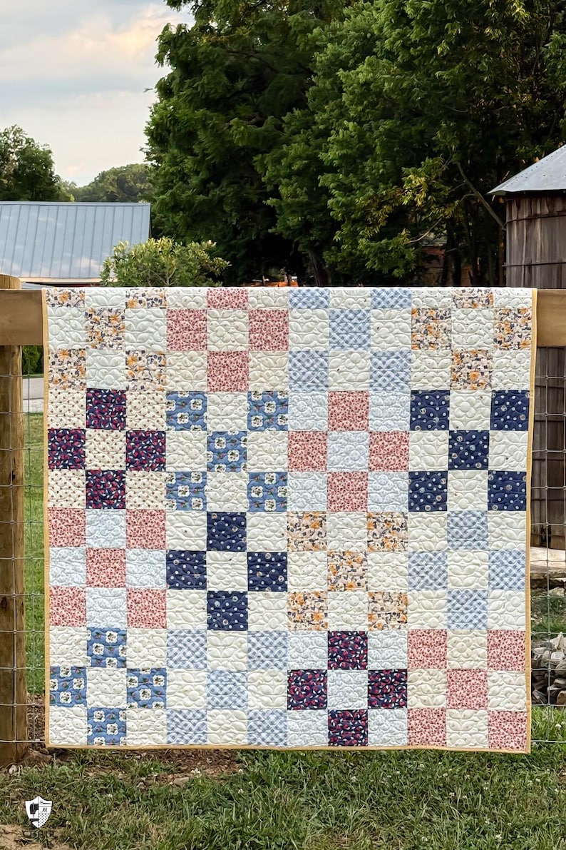 May include: A handmade quilt with a patchwork design. The quilt features a grid pattern of squares in various floral and solid prints, including pink, blue, and navy. The quilt is displayed outdoors, draped over a wooden fence, with a yellow border.