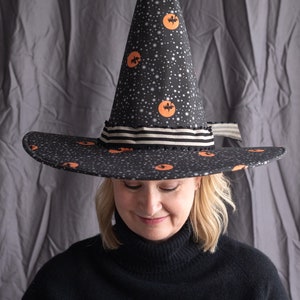 May include: A black witch hat with a pointed crown and a wide brim. The hat is decorated with orange moons and black bats on a black background with white stars. The hat has a black and white striped ribbon around the base of the crown.