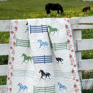 May include: A white quilt with a horse design, draped over a white fence. The quilt features horse silhouettes in green, blue, and black, with blue and green fence designs. The border is decorated with red and pink roses.