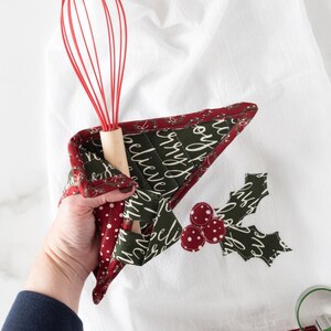 May include: A white kitchen towel with a red and green Christmas-themed design. The towel has a red border with the text "Merry Joy" and a green holly leaf design with red berries. A red whisk is tucked into the towel.