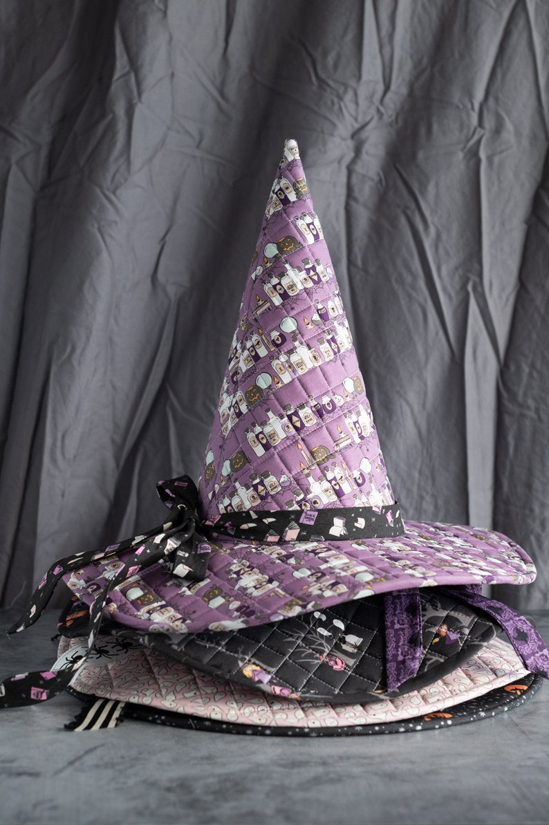May include: A stack of four purple, black, and pink witch hats with a potion and spellbook print. The top hat is tall and pointed with a wide brim.