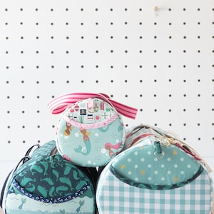 May include: Three teal and green fabric zippered bags with different patterns. The top bag has a pink and white striped strap and a mermaid pattern. The middle bag has a whale pattern. The bottom bag has a white and teal gingham pattern.