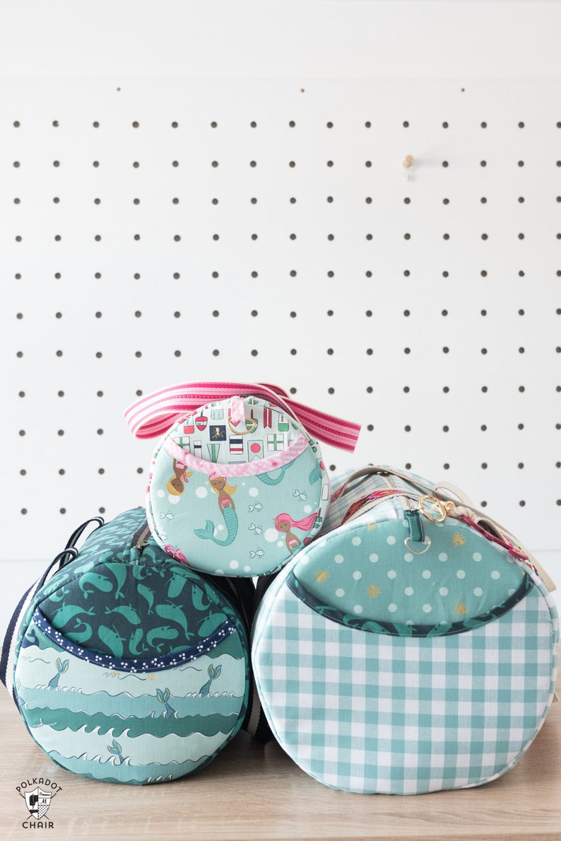 May include: Three teal and green fabric zippered bags with different patterns. The top bag has a pink and white striped strap and a mermaid pattern. The middle bag has a whale pattern. The bottom bag has a white and teal gingham pattern.