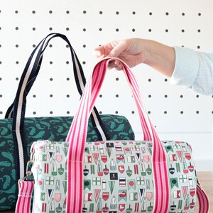 May include: Two canvas duffel bags with pink and white striped straps. The bag on the left has a green whale print. The bag on the right has a nautical flag print.