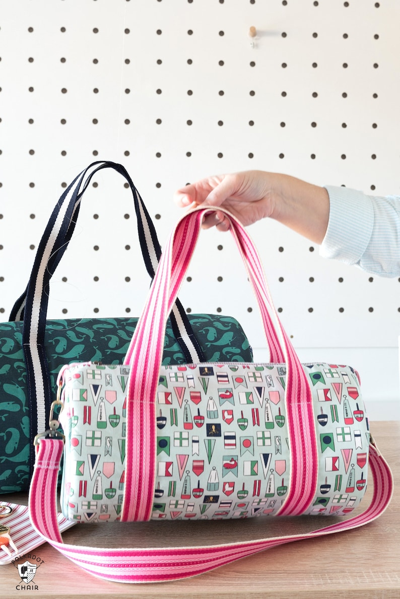 May include: Two canvas duffel bags with pink and white striped straps. The bag on the left has a green whale print. The bag on the right has a nautical flag print.