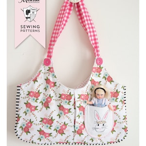 May include: A pink and white gingham fabric tote bag with a floral print and a pocket featuring a white bunny with a pink bow. The bag has a white doll inside the pocket. The text "THE RUBY LOU BAG" is printed below the bag.