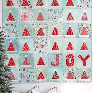 May include: A Christmas quilt with a patchwork design featuring red and white Santa hats on a light blue background. The quilt is hanging on a white wall with two white pillows with red text that says "Naughty" and "Nice".