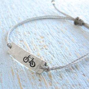 May include: A gray braided cord bracelet with a silver charm featuring a bicycle silhouette.