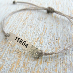 Pode incluir: Um bracelete de corda cinzenta com uma etiqueta prateada que diz "11964".
